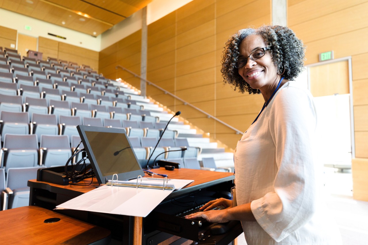 1216637043 The cheerful, mature adult female university professor pauses in her class preparation to smile for the camera. She is using the audio visual equipment.