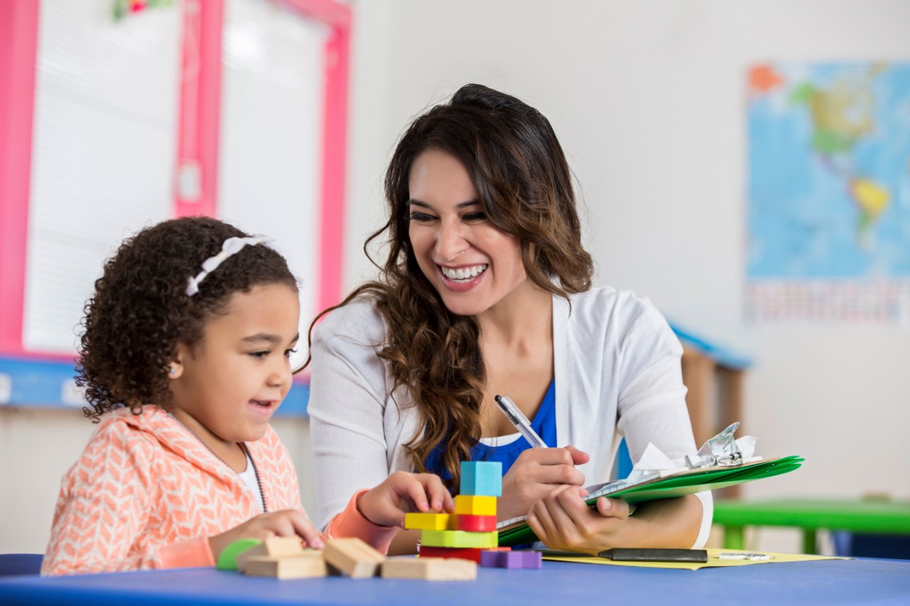 602290480 Smiling Hispanic young adult female teacher working with adorable African American little girl during preschool class. They are sitting at a table with colorful blocks. The teacher takes notes as the little girl puts the blocks together.