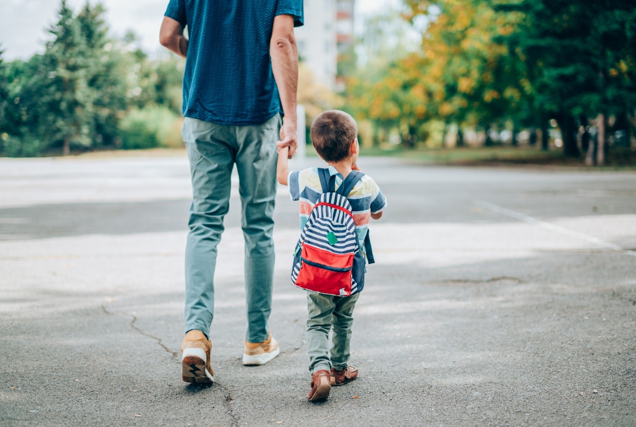 1220581522 Rear view of father who leads a little boy hand in hand to kindergarten. Father and son with backpack walking in schoolyard.