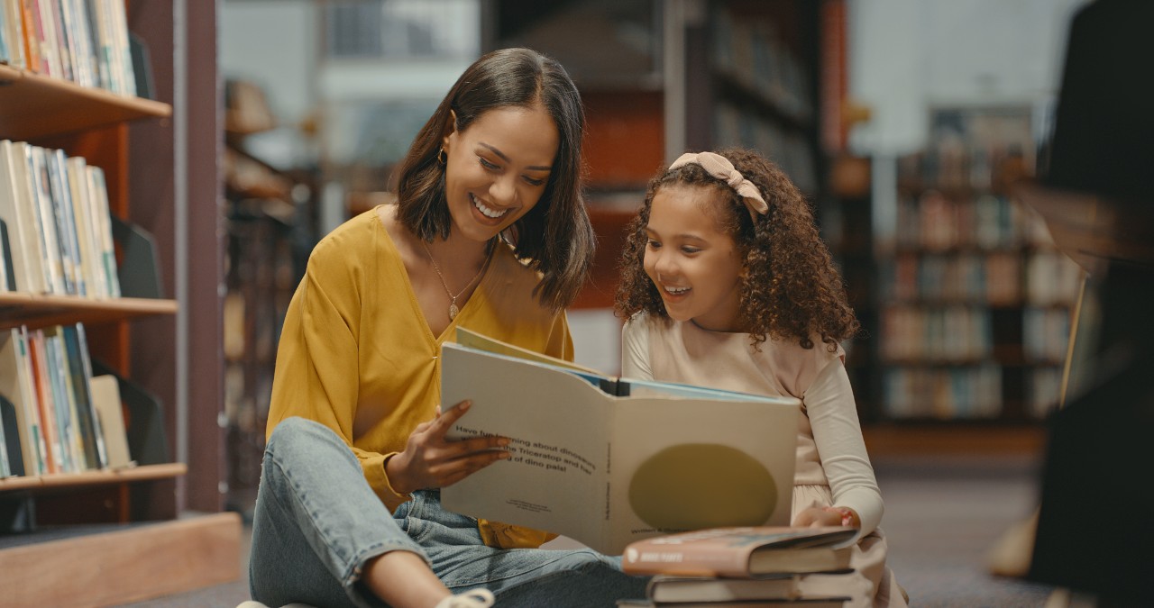 1401178460 Teacher helping a young student with her homework in the library after school. Two females are reading a book together in the bookstore. They are doing research for a project